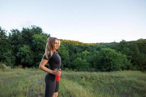 un' giovane bellissimo ragazza nel un' nero stretto completo da uomo con un' bottiglia di acqua nel sua mani. atletico ragazza nel un' stretto uniforme Lavorando all'aperto nel il parco. foto