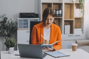 bellissimo sorridente asiatico donna d'affari Tenere un' caffè tazza mentre assunzione Appunti a ufficio. foto