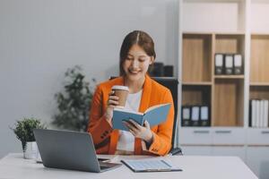 bellissimo sorridente asiatico donna d'affari Tenere un' caffè tazza mentre assunzione Appunti a ufficio. foto