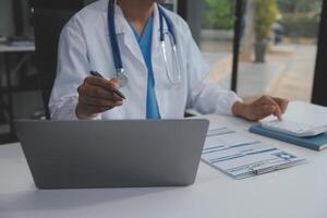 un' professionale e focalizzata asiatico femmina medico nel scrub è Lavorando e lettura medico ricerca su sua il computer portatile nel sua ufficio a un' Ospedale. foto