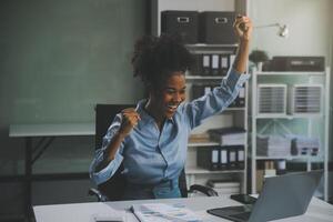 felice eccitata imprenditrice asiatica di successo che trionfa con uno smartphone per computer portatile nell'ufficio del posto di lavoro foto