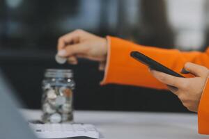 bellissimo asiatico donna d'affari inserire monete nel un' perno porcellino banca su il tavolo. Salvataggio i soldi, investimento, moneta concetto foto