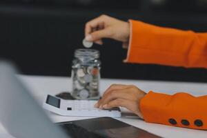 bellissimo asiatico donna d'affari inserire monete nel un' perno porcellino banca su il tavolo. Salvataggio i soldi, investimento, moneta concetto foto