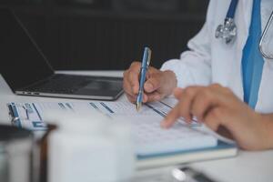 un' professionale e focalizzata asiatico femmina medico nel scrub è Lavorando e lettura medico ricerca su sua il computer portatile nel sua ufficio a un' Ospedale. foto