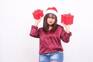 bellissimo giovane asiatico indonesiano ragazza trasporto regalo scatola a Natale Santa Claus cappello moderno rosso camicia attrezzatura sollevamento scatola su su bianca sfondo per promozione e pubblicità foto