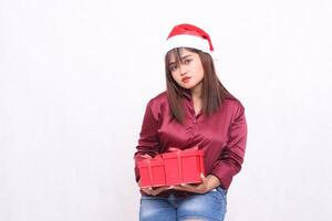 bellissimo giovane asiatico indonesiano ragazza trasporto regalo scatola a Natale Santa Claus cappello moderno brillante rosso camicia attrezzatura guardare a telecamera pianura inclinata su bianca sfondo per promozione e pubblicità foto