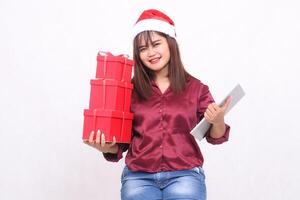 bellissimo giovane sud-est asiatico donna felicemente trasporto 3 regalo scatole ostacolare e il computer portatile tavoletta a Natale indossare Santa cappello moderno rosso camicia attrezzatura bianca sfondo per promozione e pubblicità foto