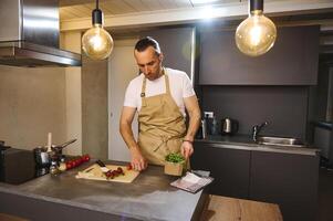 bello giovane adulto uomo anni 40, vestito nel beige capocuoco grembiule Tenere un' cartone gabbia con fresco verde le foglie di rucola, cucinando salutare cena nel il casa cucina. foto