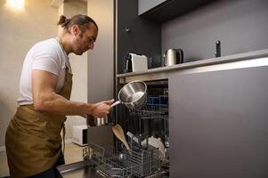 bello caucasico giovane uomo nel beige capocuoco grembiule, prende su un' pulito casseruola a partire dal un sovraccarico automatico lavastoviglie con pulito sterilizzare cucina utensili. moderno cucina elettrodomestici, stile di vita Visualizza foto