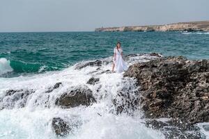 un' donna sta su un' roccia nel il mare durante un' tempesta. vestito nel un' bianca lungo vestire, il onde rompere su il rocce e bianca spray si alza. foto