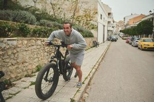 europeo giovane uomo spingendo il suo elettrico montagna bicicletta su il strada. attivo stile di vita. sport. avventura viaggio su bicicletta. utilizzando el-bicicletta anno Domini ecologicamente amichevole modalità di urbano trasporto foto
