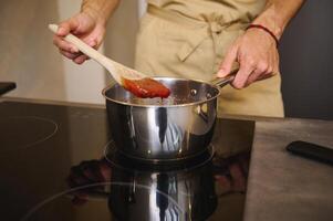 avvicinamento capocuoco in piedi di elettrico induzione fornello, utilizzando di legno mestolo, eccitante bollente pomodoro succo, cucinando fatti in casa passata a partire dal biologico succoso pomodori nel il cucina foto