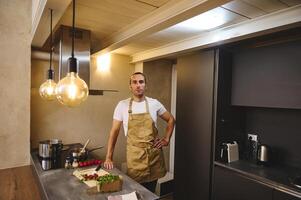 autentico ritratto di un' bello giovane adulto uomo nel beige dello chef grembiule, in piedi a cucina contatore e guardare a telecamera mentre cucinando cena, nel il moderno interno di minimalista casa cucina foto