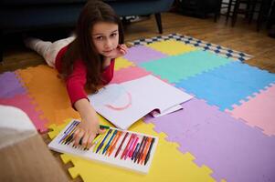 poco ragazza sorridente guardare a telecamera mentre assunzione un' colorato matita su di un' scatola con pastello pastelli, dire bugie su sua pancia su un' multicolore puzzle tappeto. le persone. formazione scolastica. indietro per scuola concetto foto