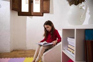 autentico ritratto erudito bambino ragazza, elementare età scolara lettura un' libro nel sua camera. nelle vicinanze scaffale con libri e scuola forniture. Multi colorato puzzle tappeto su il pavimento. autismo concetto foto