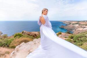 bionda con lungo capelli su un' soleggiato riva del mare nel un' bianca fluente vestire, posteriore Visualizza, seta tessuto agitando nel il vento. contro il fondale di il blu cielo e montagne su il spiaggia. foto