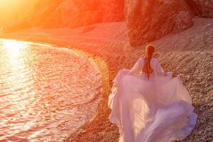 misterioso donna silhouette lungo capelli passeggiate su il spiaggia oceano acqua, mare ninfa vento ascolta per il onda. lanci su un' lungo bianca vestire, un' divine tramonto. artistico foto a partire dal il indietro senza un' viso