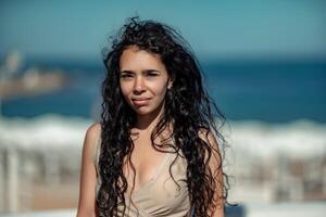 mare donna riposo. ritratto di un' donna con lungo Riccio nero capelli nel un' beige vestito sta su un' balcone contro il fondale di il mare. turista viaggio per il mare. foto