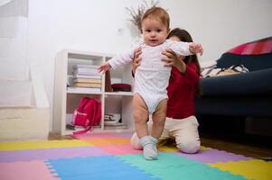 selettivo messa a fuoco su adorabile bambino ragazzo provando per il suo primo passaggi su il colorato puzzle tappeto con il Aiuto di il suo giovane amorevole cura sorella. bambini crescita e sviluppo. infanzia. infanzia. famiglia foto