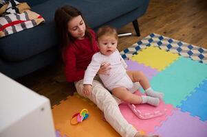 adorabile bello sorella Tenere supporto sua minore fratello, un' carino bambino ragazzo provando per fare il suo primo passi, seduta su un' Multi colorato puzzle tappeto a casa. bambini. crescita. famiglia relazioni foto