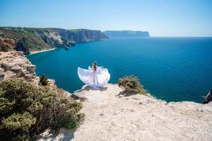 bionda con lungo capelli su un' soleggiato riva del mare nel un' bianca fluente vestire, posteriore Visualizza, seta tessuto agitando nel il vento. contro il fondale di il blu cielo e montagne su il spiaggia. foto