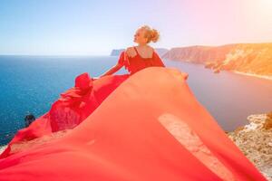 donna mare rosso vestire. bionda con lungo capelli su un' soleggiato riva del mare nel un' rosso fluente vestire, indietro Visualizza, seta tessuto agitando nel il vento. contro il fondale di il blu cielo e montagne su il spiaggia. foto