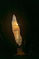 un' grotta con un' piccolo apertura nel esso e il sole splendente attraverso esso. il sole è ambientazione e il acqua è calma. foto