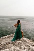 donna mare viaggio verde vestire. lato Visualizza un' contento donna con lungo capelli nel un' lungo menta vestito in posa su un' spiaggia con calma mare bokeh luci su soleggiato giorno. ragazza su il natura su blu cielo sfondo. foto