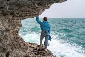 un' donna nel un' blu giacca sta su un' roccia sopra un' scogliera sopra il mare e sembra a il furioso oceano. ragazza viaggiatore riposa, pensa, sogni, gode natura. pace e calma paesaggio, ventoso tempo atmosferico. foto