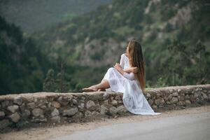 un' donna nel un' bianca vestito è seduta su un' pietra parete prospiciente un' montagna. il scena è sereno e tranquillo, calmo, con il donna di lungo capelli soffiaggio nel il vento. concetto di calma e la tranquillità. foto