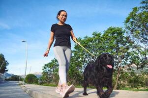 Visualizza a partire dal il parte inferiore di un' contento giovane donna nel gli sport Abiti, a piedi sua cane su guinzaglio e godendo mattina correre con sua animale domestico nel il montagne natura foto