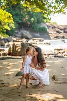 giovane donna madre con un' poco figlia nel bianca vestiti su riva del mare nel il ombra di alberi e palme foto