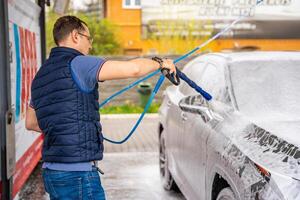 giovane uomo lavaggi il suo auto a un' Fai da te auto lavare utilizzando un' tubo flessibile con pressurizzato acqua e schiuma. alto qualità foto