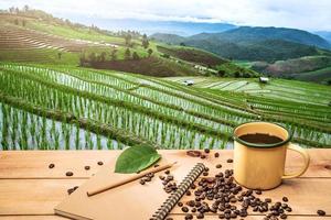 tazza di caffè caldo con chicchi di caffè biologico sul tavolo di legno sopra il paesaggio delle montagne con la luce del sole e bellissime risaie foto