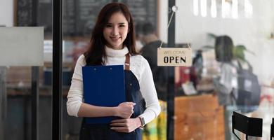 ritratto di una bella donna asiatica in un grembiule in piedi in una caffetteria, possiede una caffetteria, il concetto di un'attività di cibo e bevande. gestione del negozio da parte di una donna d'affari. foto