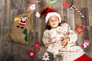 adorabile bambina che indossa un cappello da Babbo Natale sdraiato sul pavimento di legno foto