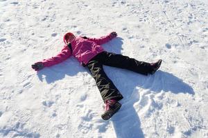 bambina che fa un angelo di neve indossando vestiti da neve foto