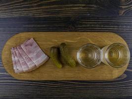 due bicchieri appannati con vodka fredda su una tavola di legno con pancetta e cetrioli sottaceto. spuntino tradizionale russo foto