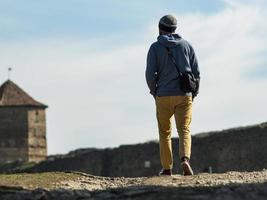un uomo con un maglione con cappuccio e un cappello lavorato a maglia sta camminando lungo la strada per la fortezza sullo sfondo della torre e del cielo blu foto