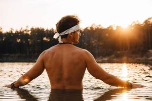 uomo con muscoli perfetti sta in acqua al tramonto lanciando le mani di lato foto