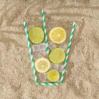 creativo estate composizione con Limone e lime fette, potabile cannuccia e ghiaccio cubi su sabbioso sfondo. minimo bevanda concetto. superiore giù limonata disposizione. esotico estate spiaggia festa idea. piatto posizione. foto