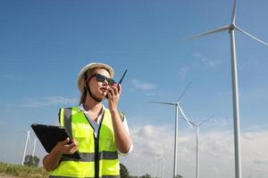 il fiducioso donna nel bianca casco Lavorando con digitale tavoletta e utilizzando walkie talkie a un' rinnovabile energia azienda agricola. femmina ispettore controllo il funzionamento di vento turbine all'aperto foto