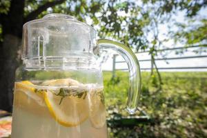 limonata fatta in casa a base di limoni in una grande brocca di vetro sul tavolo in giardino. una brocca con limone e menta si erge per strada sullo sfondo del verde in una calda giornata estiva. foto