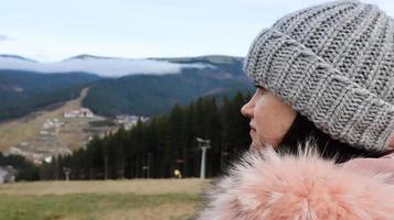 vista posteriore di una turista di montagna donna in una giacca rosa con pelliccia e un cappello a maglia grigio su uno sfondo di montagne che guardano nel paesaggio autunnale della foresta e del cielo nuvoloso. foto