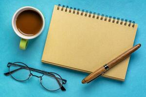 vuoto spirale taccuino con lettura occhiali, penna e caffè su blu carta sfondo foto