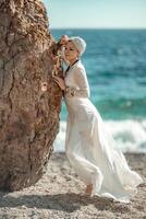 mezzo anziano donna sembra bene con biondo capelli, boho stile nel bianca lungo vestito su il spiaggia decorazioni su sua collo e braccia. foto