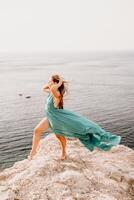 donna mare verde vestire. lato Visualizza un' contento donna con lungo capelli nel un' lungo menta vestito in posa su un' spiaggia con calma mare bokeh luci su soleggiato giorno. ragazza su il natura su blu cielo sfondo. foto