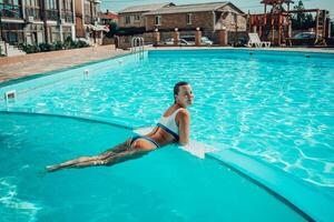 donna nuoto piscina. contento donna con bagnato capelli e elegante occhiali da sole prendere il sole nel piscina foto