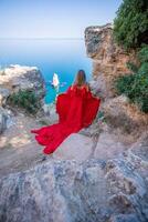 rosso vestito mare donna. contento donna con fluente capelli nel un' lungo fluente rosso vestito sta su un' roccia vicino il mare. viaggio concetto, foto sessione a mare