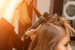 il parrucchiere prepara un' donna, fa un' acconciatura con riccioli utilizzando un' arricciatura ferro da stiro. lungo leggero Marrone naturale capelli. se stesso cura concetto. foto